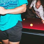 Man wearing black lounge shorts playing pool with a man behind in playing pool wearing light linen hoodie.