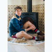 Man sitting in room next to fireplace wearing jacket, red underwear, and chunky knit stripe socks.