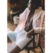Man sitting down in chair holding coffee mug wearing open light pink dress shirt and color block blues slim fit boxers.