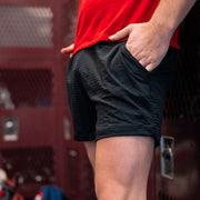 Close up of man in locker room wearing black lounge shorts and red shirt with his hand in pocket.