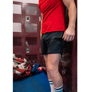 Man in locker room wearing red shirt, black lounge shorts, and black/blue stripe crew socks.