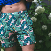 Man holding shirt up wearing pink palms lounge shorts standing in front of a bush.