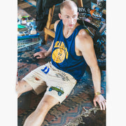 Man sitting on rug next to motorbike wearing Beige lounge short that says "cash rules everything around me" with various icons like 11 patch, truck, football, skull and bones etc. 