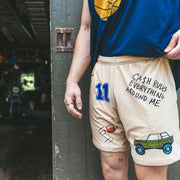 Man standing in doorway wearing blue basketball shirt and Beige lounge short that says "cash rules everything around me" with various icons like 11 patch, truck, football, skull and bones etc. 