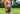 Shirtless man standing in forest wearing hiking backpack and olive green lounge shorts.