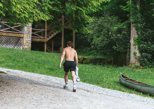 Shirtless man walking in forest with canoe and cabin in background wearing black lounge shorts.