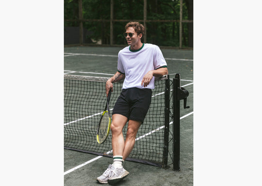 Man leaning on tennis court wearing white t-shirt and black lounge shorts.