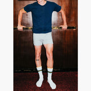 Man standing against wood background wearing navy shirt, grey slim fit boxers and striped Amalfi athletic crew socks.
