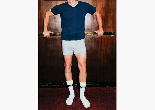 Man standing against wood background wearing navy shirt, grey slim fit boxers and striped Amalfi athletic crew socks.
