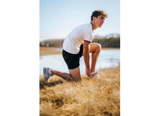 Man in field adjusting shoe wearing shite shirt and black shorts.