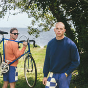 Man in blue sweater and beach patrol lounge shorts on the grass next to water with friend holding a bike in the background.