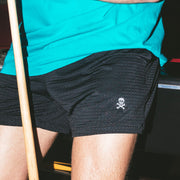 Man wearing black mesh shots with white skull leaning against pool table.