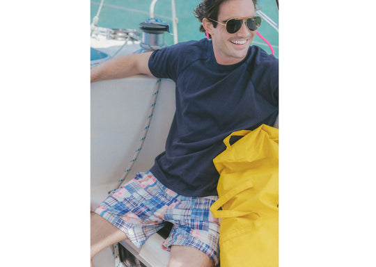 Man sitting on boat smiling wearing blue shirt and blue madras print lounge shorts.