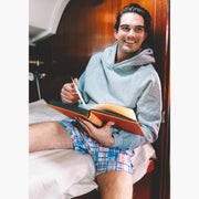 Man sitting on bed reading book wearing grey sweatshirt and blue madras slim fit boxers.