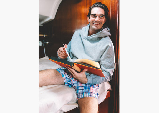Man sitting on bed reading book wearing grey sweatshirt and blue madras slim fit boxers.