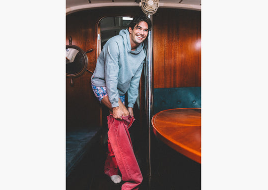Man on boat stepping into red jeans and also wearing grey sweatshirt and blue madras slim fit boxers