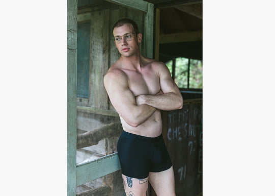 Shirtless man with arms crossed standing in front of open cabin door wearing black boxer briefs.