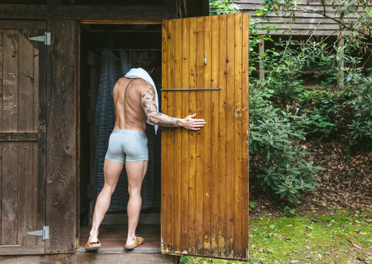 Back shot of a shirtless man walking into outdoor shower with towel on shoulder wearing grey boxer briefs.