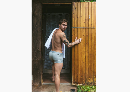 Back shot of shirtless man walking into outdoor shower with towel on shoulder wearing grey boxer briefs.
