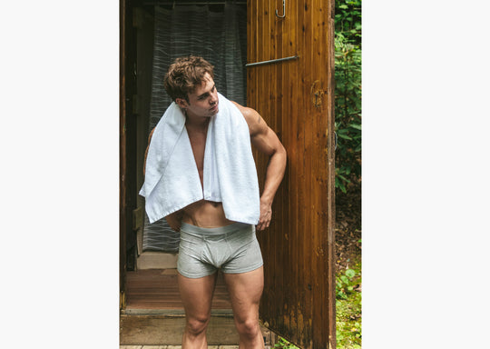 Shirtless man standing outside of outdoor shower with towel on shoulder wearing grey boxer briefs.