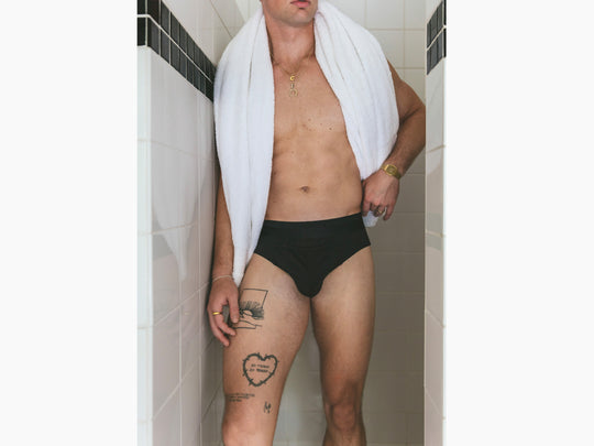 Shirtless man standing in shower wearing gold chain, towel and black briefs.