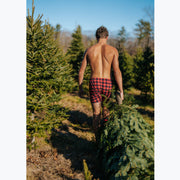 Back shot of a shirtless man walking in forest wearing red/black buffalo plaid slim fit boxers. he is pulling a tree behind him. 