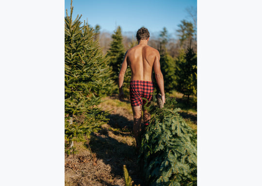 Back shot of a shirtless man walking in forest wearing red/black buffalo plaid slim fit boxers. he is pulling a tree behind him. 