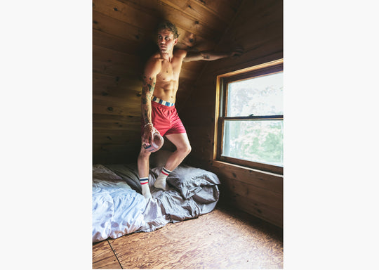Shirtless man standing on bed holding a football wearing burgundy lounge shorts and navy/red crew socks.