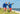 Two men walking together at the beach both wearing blue tops and red/white beach patrol lounge shorts.