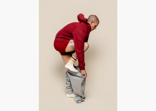 Man standing in studio putting on grey sweat pants and also wearing burgundy boxer briefs and oodie.