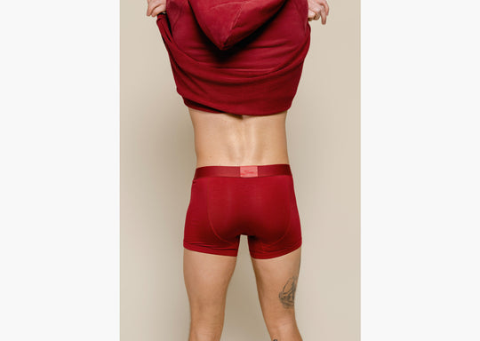 Backshot of standing in studio taking off burgundy sweatshirt and wearing burgundy boxer briefs. 