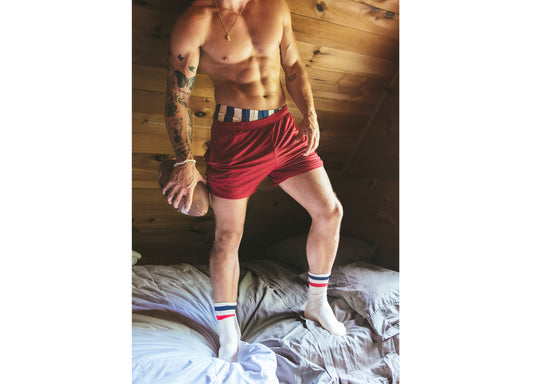 Man standing on bed shirtless holding a football and wearing burgundy mesh lounge shorts.
