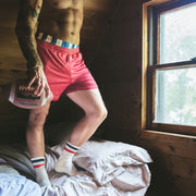 Man standing on bed shirtless holding a football and wearing burgundy mesh lounge shorts.