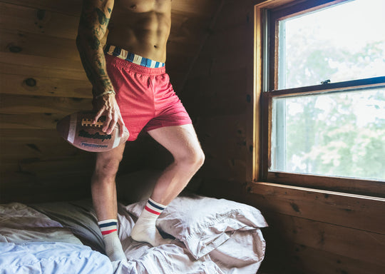 Man standing on bed shirtless holding a football and wearing burgundy mesh lounge shorts.