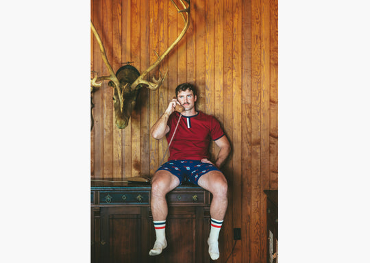 Man sitting in house with wood walls on phone wearing burgundy vintage ringer shirt