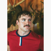 Close up of man standing in house with wood walls wearing burgundy vintage ringer shirt