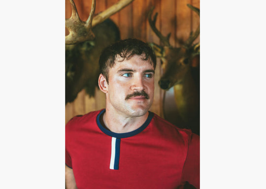 Close up of man standing in house with wood walls wearing burgundy vintage ringer shirt