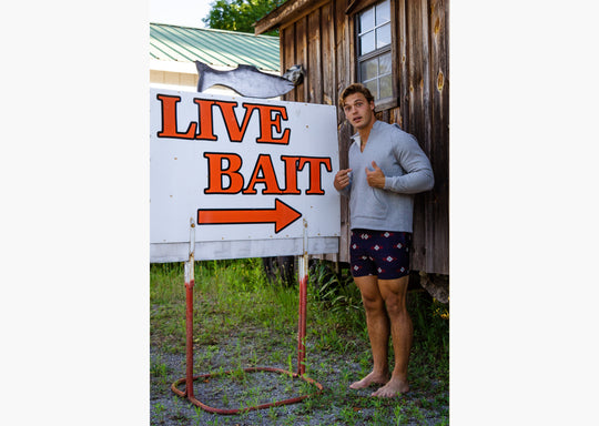 Man wearing cabin club lounge shorts and grey hoodie. He is standing next to large sigh that says live bait 