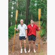 Two men standing next to each other in the woods. Both wearing cabin club lounge shorts and holding a canoe paddle. 