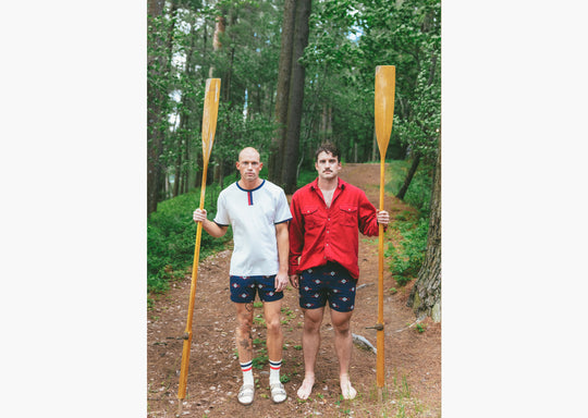 Two men standing next to each other in the woods. Both wearing cabin club lounge shorts and holding a canoe paddle. 