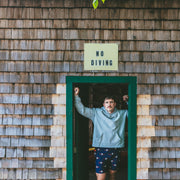 Man in grey hoodie and cabin club lounge shorts standing in doorway of cabin with No Diving sign.
