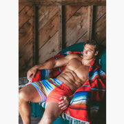 Shirtless man sitting in chair wrapped in blanket wearing Canoe Stripe lounge shorts.