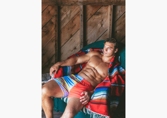 Shirtless man sitting in chair wrapped in blanket wearing Canoe Stripe lounge shorts.