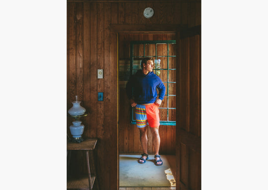 Man wearing navy sweatshirt and Canoe stripe lounge short standing inside looking out the window.