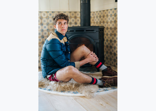 Man sitting in room next to fireplace wearing jacket, red underwear, and chunky knit stripe socks.