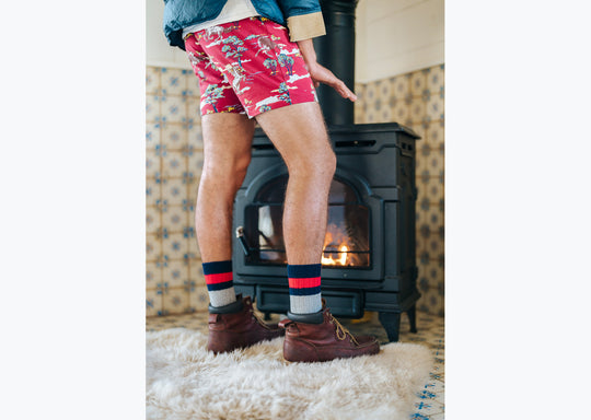 Man standing in room next to fireplace wearing jacket, red underwear, and chunky knit stripe socks.