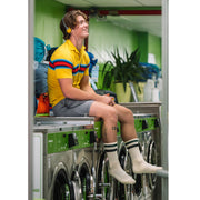 Man sitting on top of laundry machine wearing headphones, a yellow shirt, grey lounge shorts and classic crew socks in black stripe.