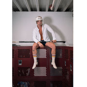 Man sitting on top of locker wearing hockey helmet, orange and black classic crew socks, and holding a hockey stick.