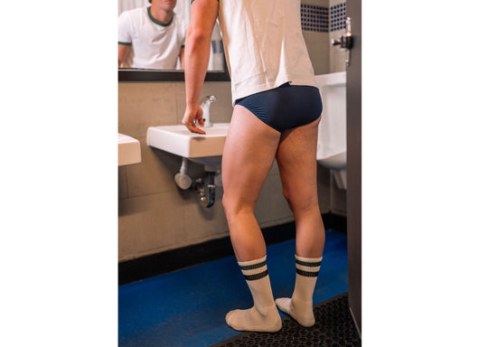 Man standing in bathroom in front of sink wearing blue boxers and black stripe classic crew socks.