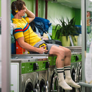 Man sitting on top of laundry machine wearing headphones, a yellow shirt, grey lounge shorts and classic crew socks in black stripe.